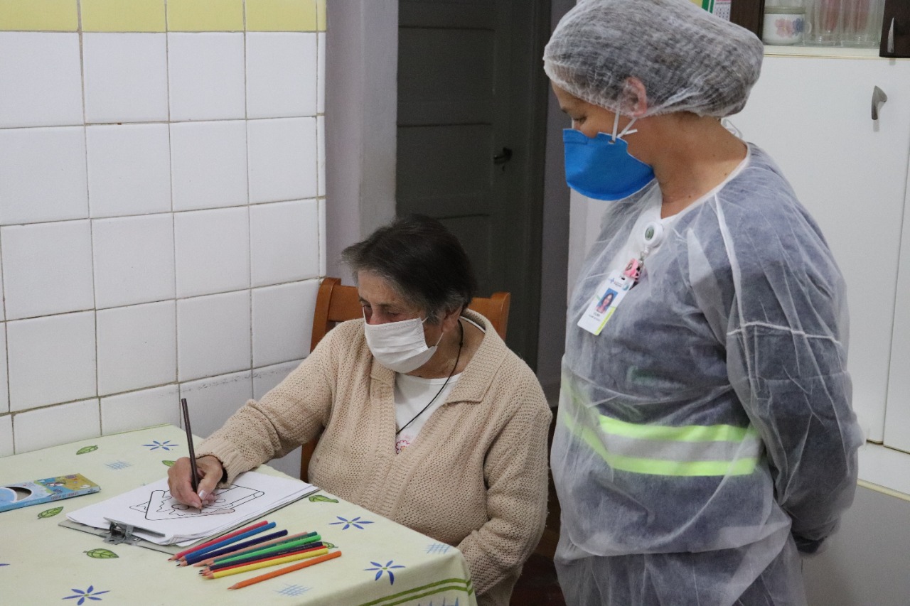 Na imagem há uma profissional da saúde em pé olhando para uma senhora que está sentada enquanto pinta um desenho que está em cima da mesa. Na mesa também está alguns lápis coloridos.